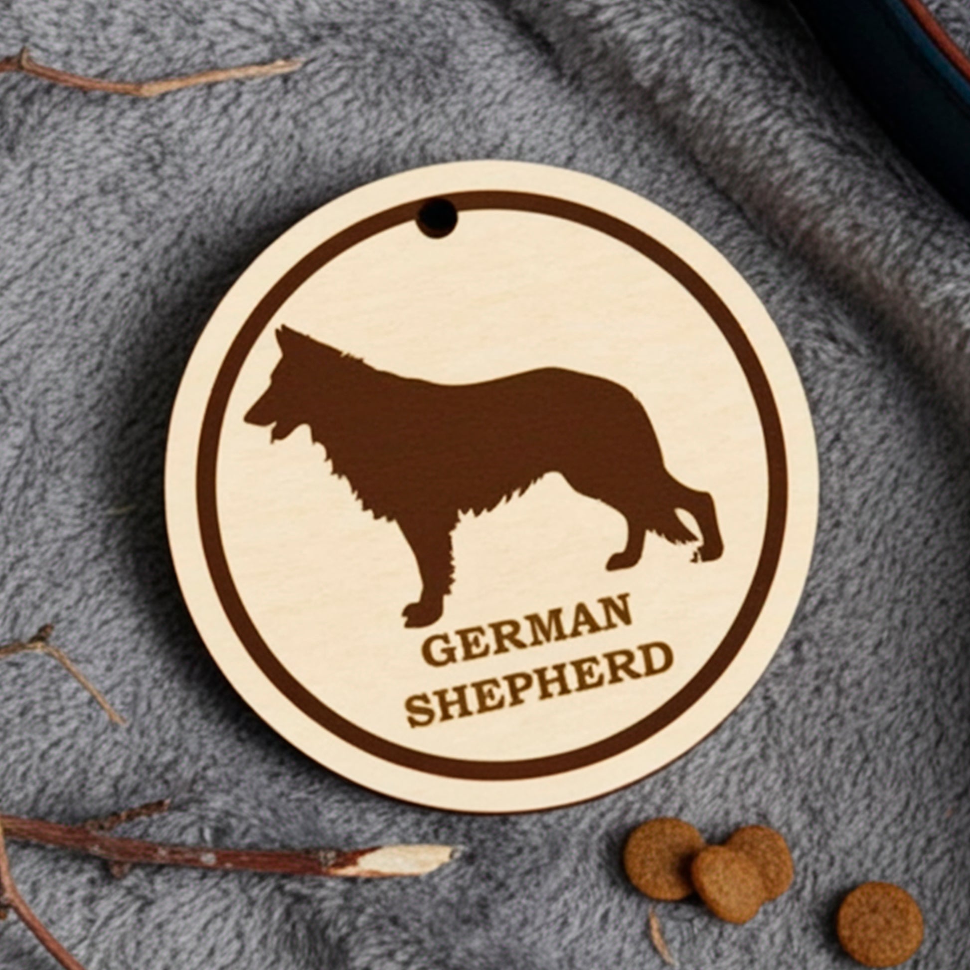 Laser cut wooden plywood dog tag  featuring an engraved black silhouette and breed name on a light wood round charm. The tag is displayed on a soft gray blanket next to a dark blue leather collar, dried sticks, and dog kibble, showing its use as a pet ID or gift