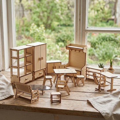 Large set of miniature doll furniture made of laser cut plywood. Includes a wardrobe, bookshelf, table, chairs, and a bench. Resting on a wooden surface by a window with garden views. Natural wood finish with visible dark burnt edges.