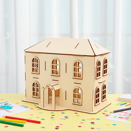 Laser cut  plywood wooden two-story dollhouse model with arched windows, a front porch, and a pitched roof, displayed on a yellow table covered in colorful paper stars and drawing supplies in a bright room