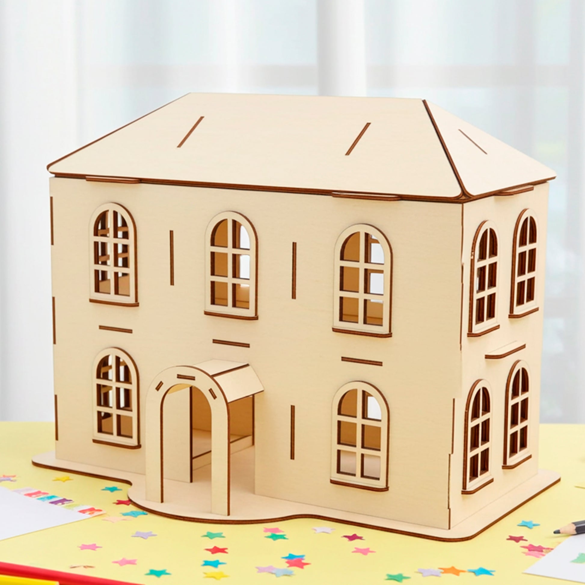 Laser cut  plywood wooden two-story dollhouse model with arched windows, a front porch, and a pitched roof, displayed on a yellow table covered in colorful paper stars and drawing supplies in a bright room