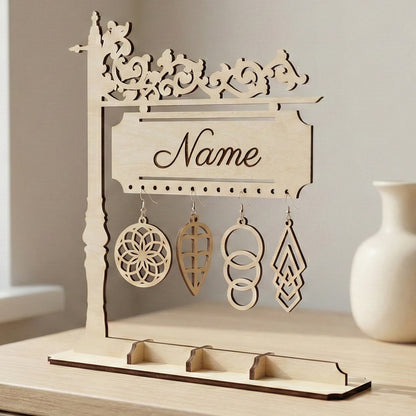 Personalized laser cut plywood earring stand on a dresser. Features an ornate scrollwork top and a sign engraved with "Name." Four different geometric wooden earrings hang from the pre-drilled holes above a jewelry tray base.