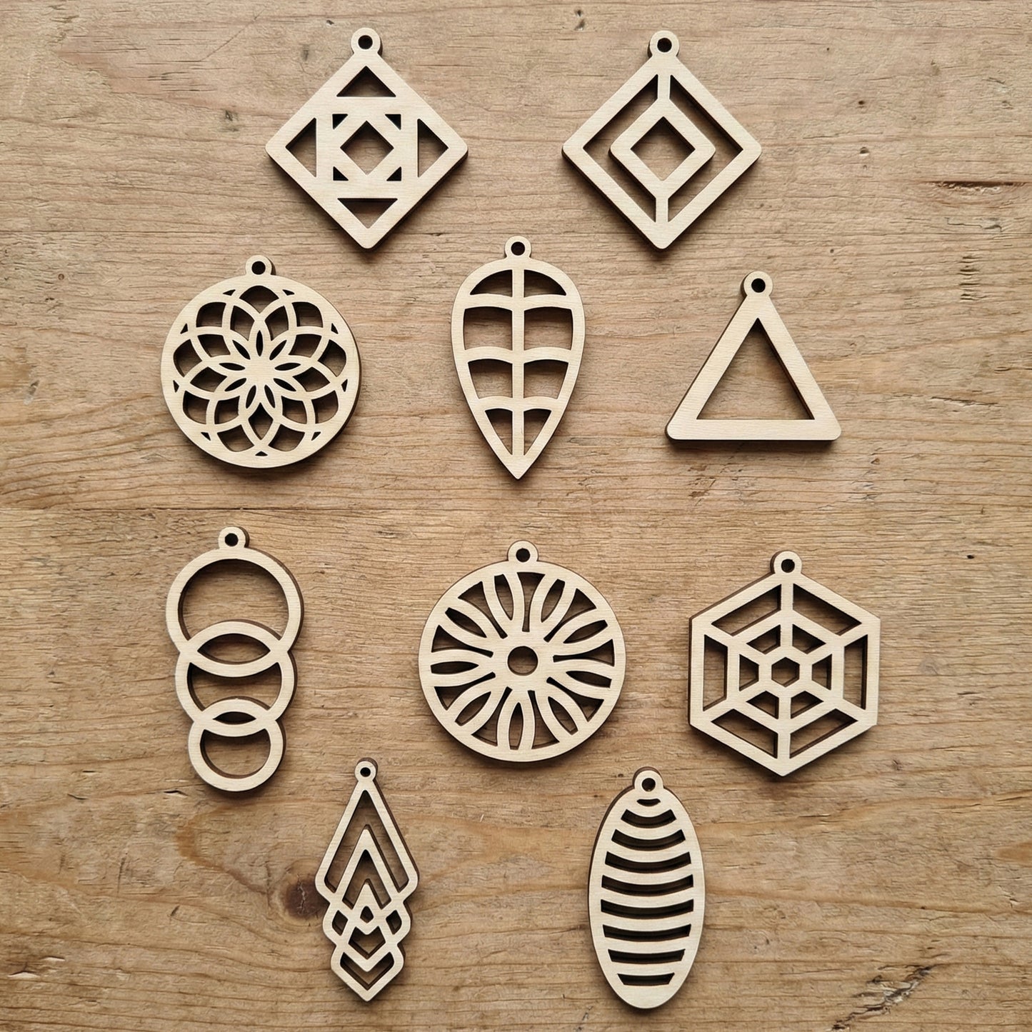 Collection of ten different geometric laser cut plywood earring designs laid flat on a wood surface. Includes circles, diamonds, and leaf shapes with intricate interior cutouts. Each piece has a small top hole for earring hooks.