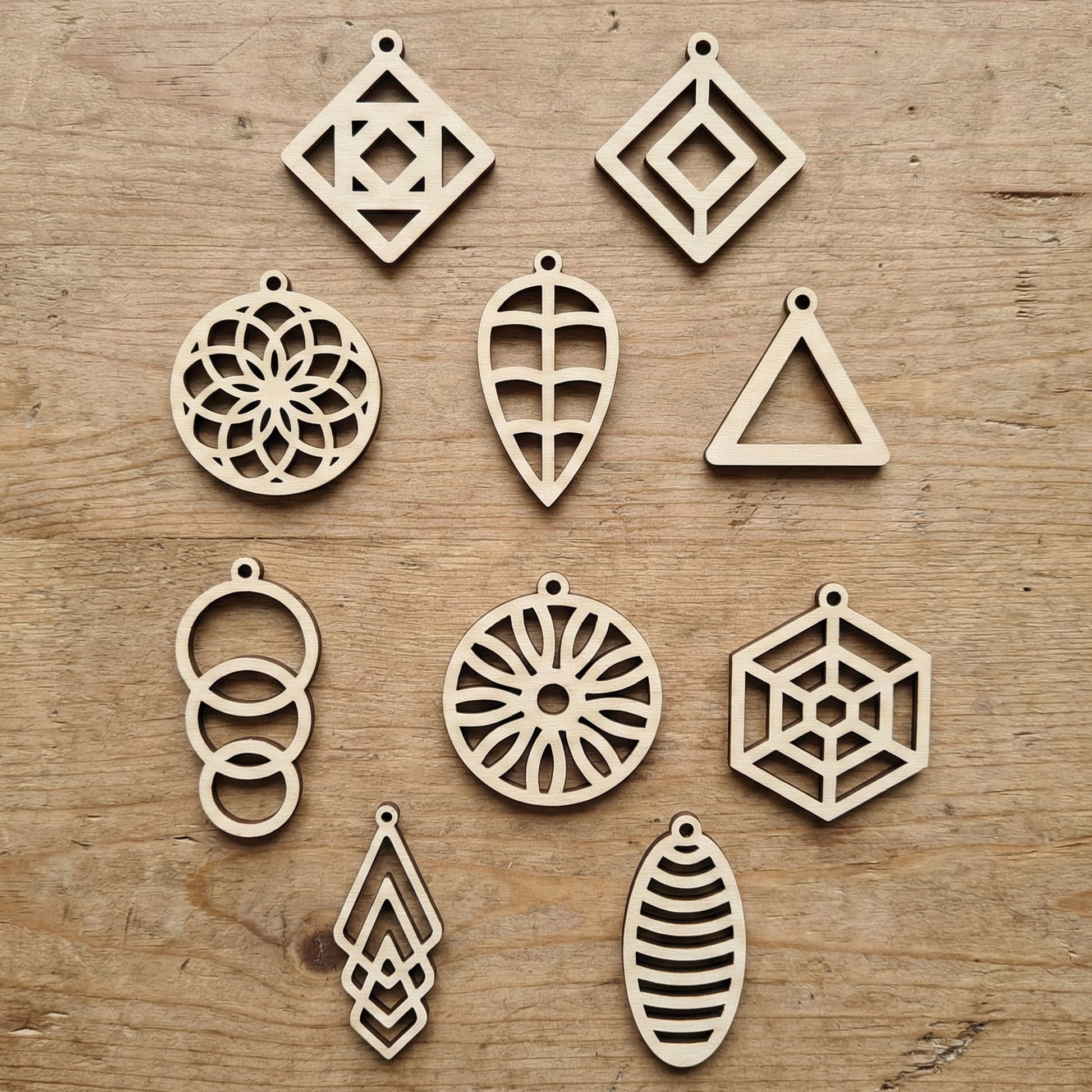 Collection of ten different geometric laser cut plywood earring designs laid flat on a wood surface. Includes circles, diamonds, and leaf shapes with intricate interior cutouts. Each piece has a small top hole for earring hooks.