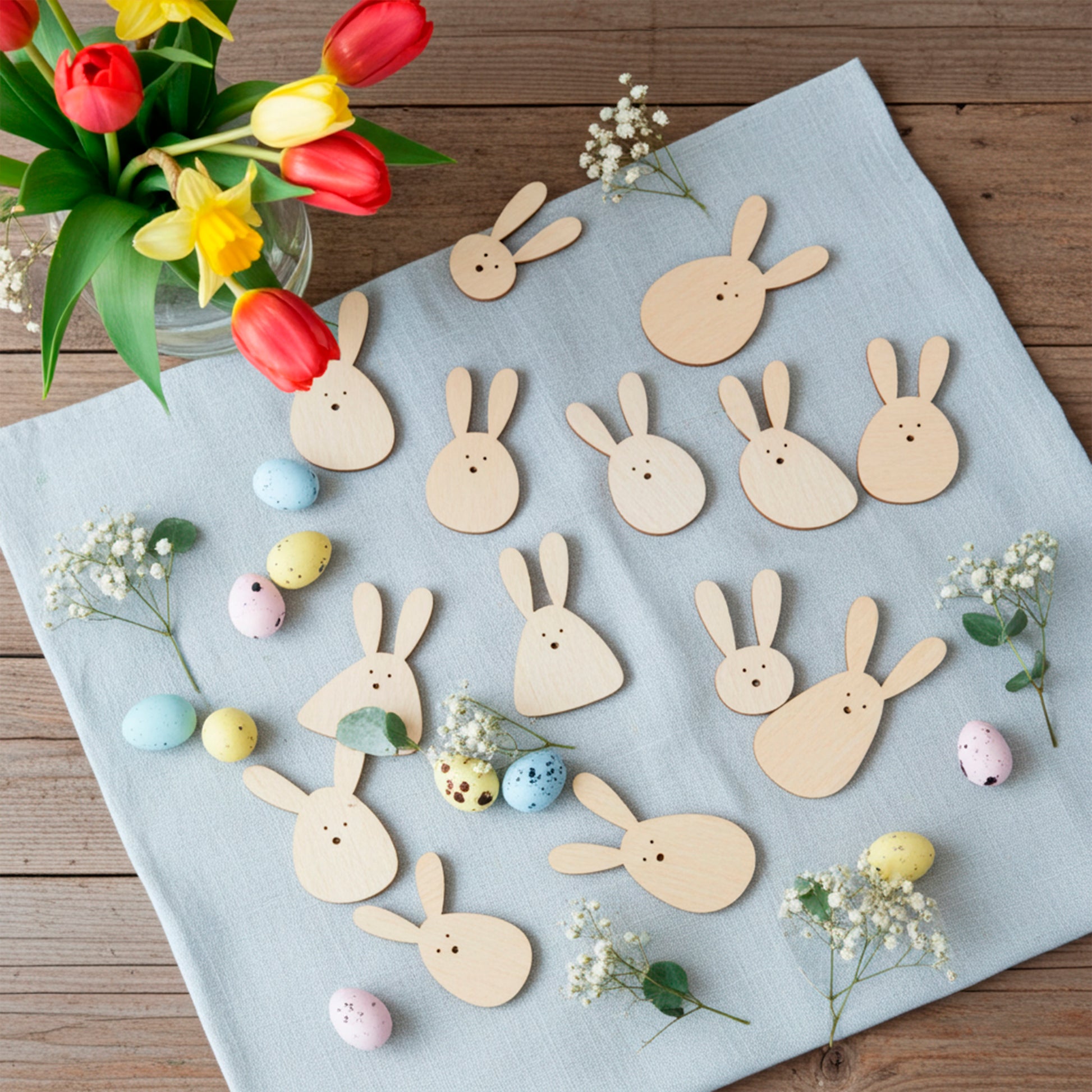 A collection of various laser cut plywood wooden Easter bunny shapes and pastel speckled eggs arranged on a light blue linen cloth, with a vase of red and yellow tulips.