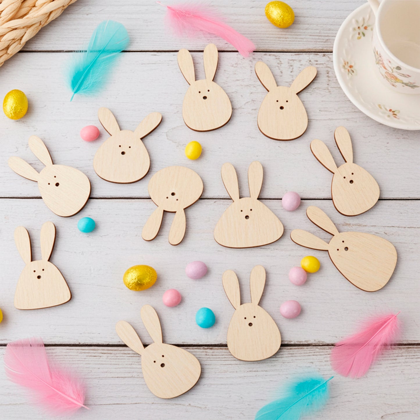 A vibrant flat lay of multiple small laser cut plywood wooden Easter bunny cutouts, arranged with colorful candies, golden foil eggs, and pink and blue feathers on white wooden planks
