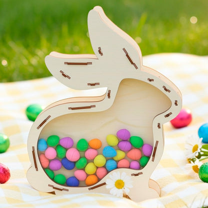 3D laser cut plywood wooden bunny shaped shaker box with a clear acrylic front, filled with small colorful candy eggs, sitting on a yellow checkered blanket in bright sunlight