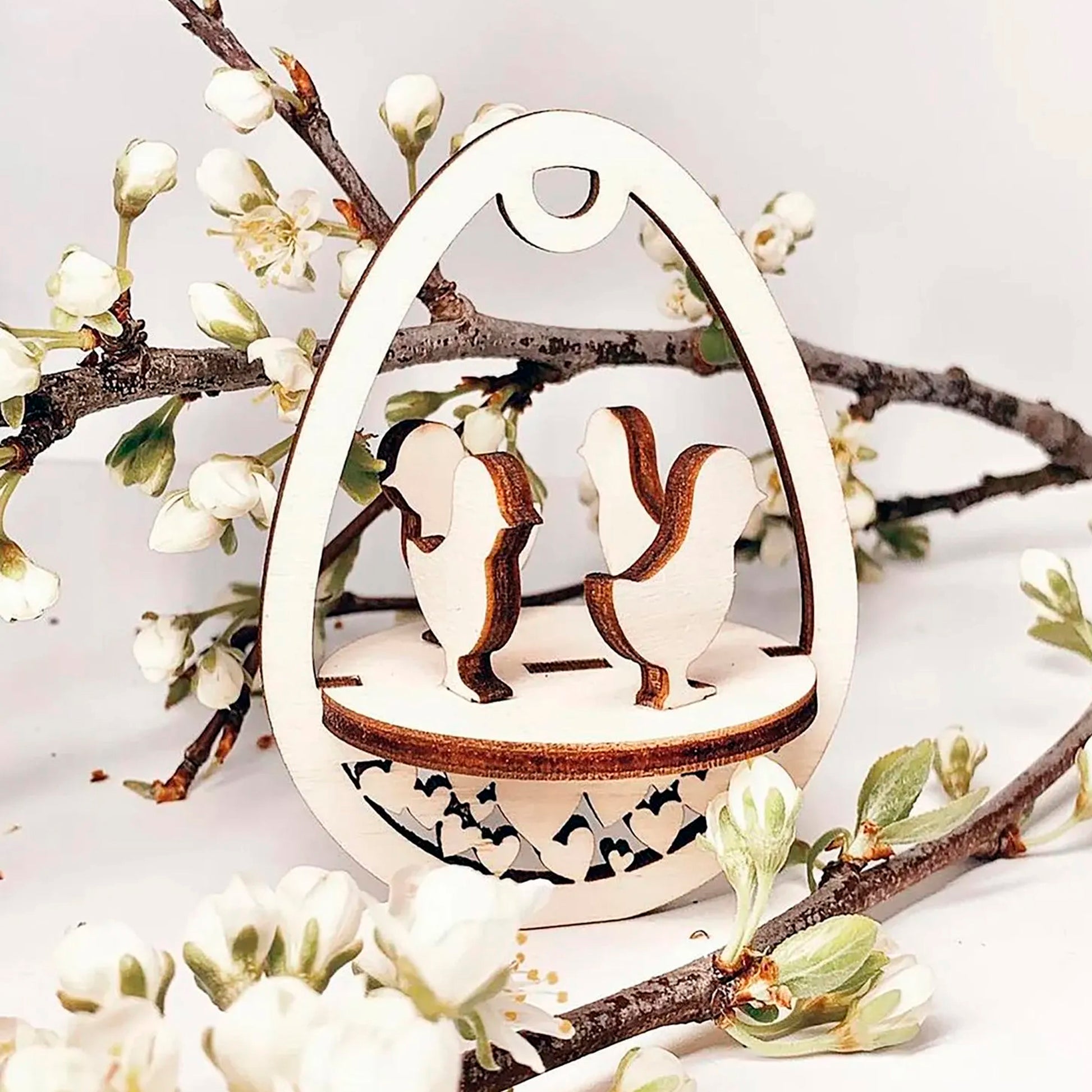 Laser cut plywood wooden Easter egg ornament featuring two chickens standing on a patterned base, framed by white blooming spring tree branches on a clean white background.