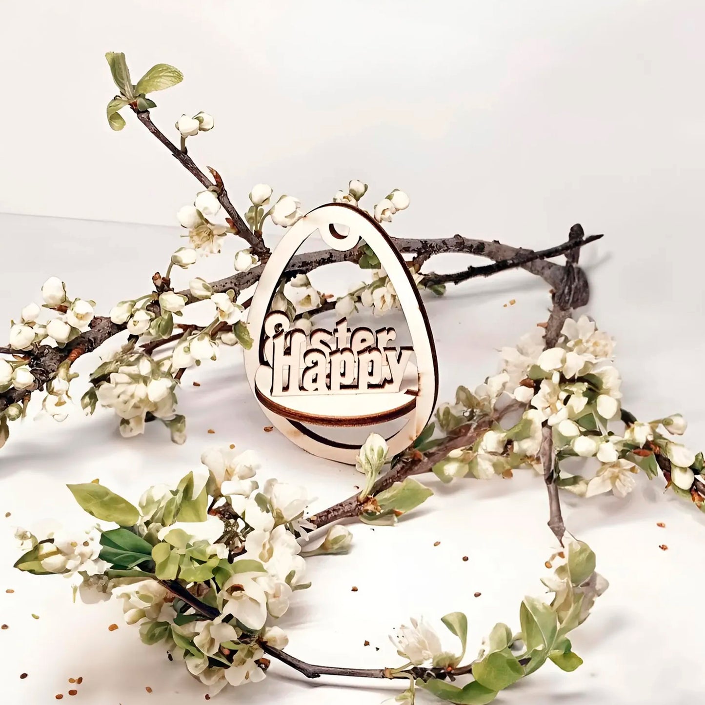Laser cut plywood wooden Easter egg ornament with the layered, etched text "Happy Easter" inside, hanging among white blooming spring tree branches on a clean white background