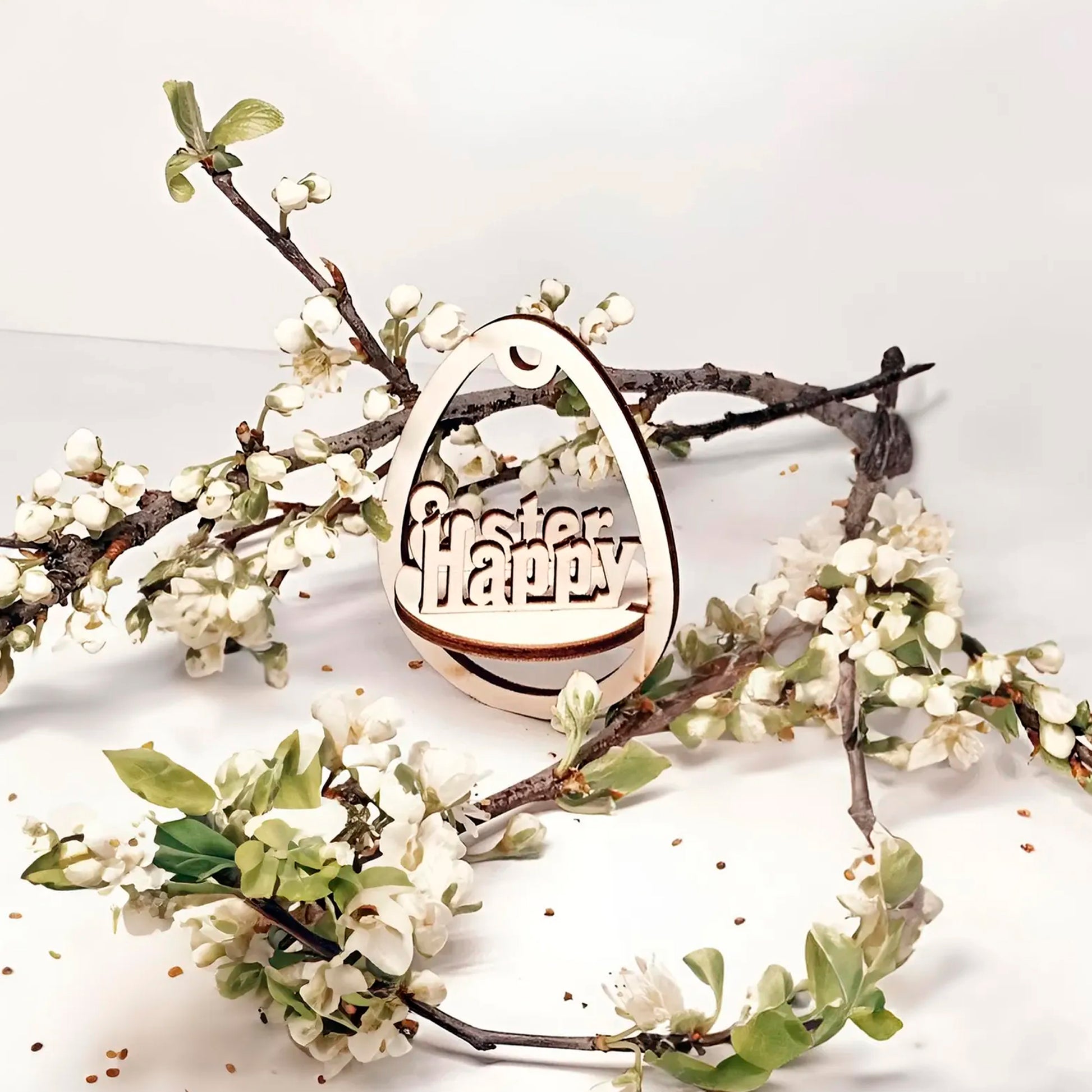 Laser cut plywood wooden Easter egg ornament with the layered, etched text "Happy Easter" inside, hanging among white blooming spring tree branches on a clean white background