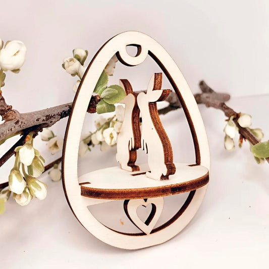 Laser cut plywood wooden Easter egg ornament featuring two kissing bunnies and a small heart on the base, framed by white blooming spring tree branches on a clean white background