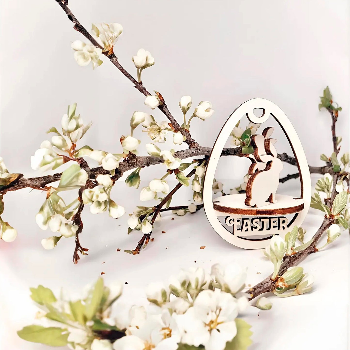 Laser cut plywood wooden Easter egg ornament with a detailed cutout of a playful bunny standing above the etched word "EASTER," framed by white blooming spring tree branches on a white background
