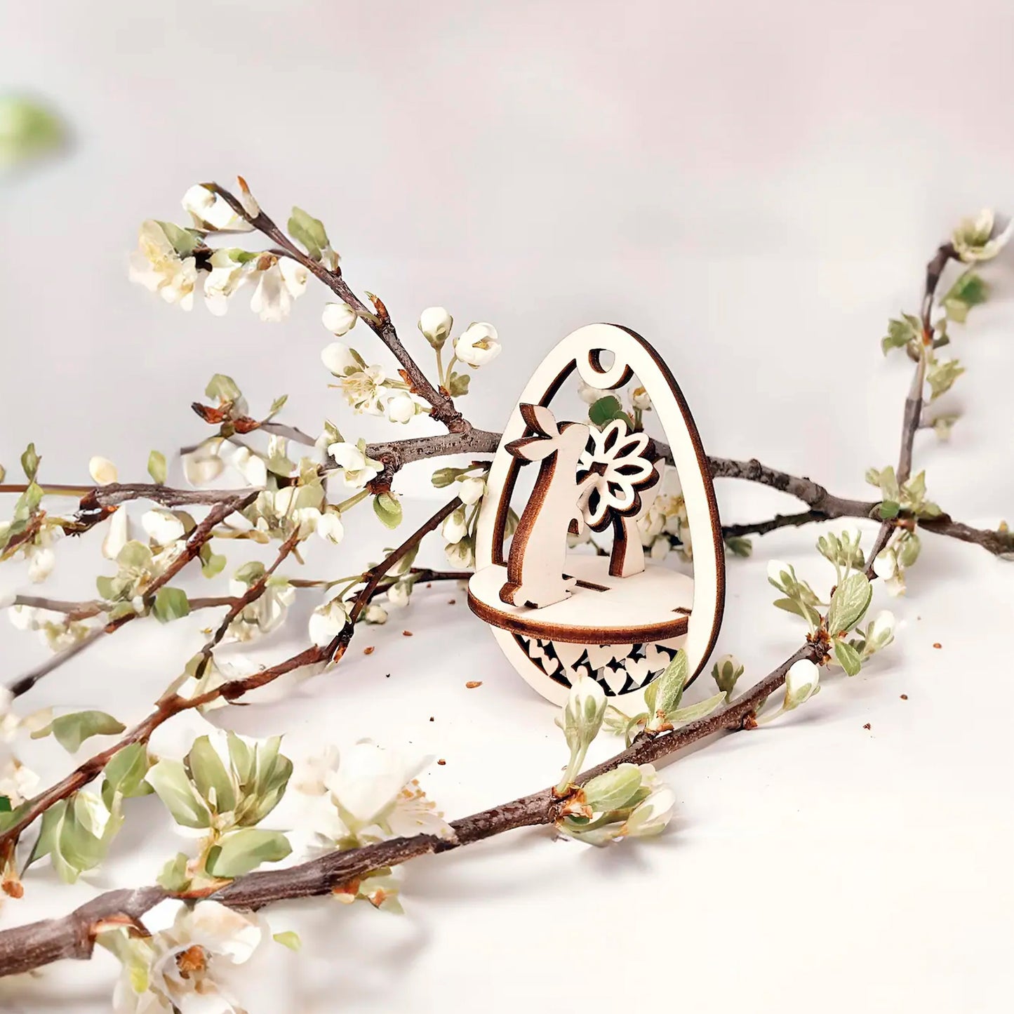 Laser cut  plywood wooden Easter egg ornament with a detailed cutout of a bunny holding a flower, framed by white blooming spring tree branches on a white background