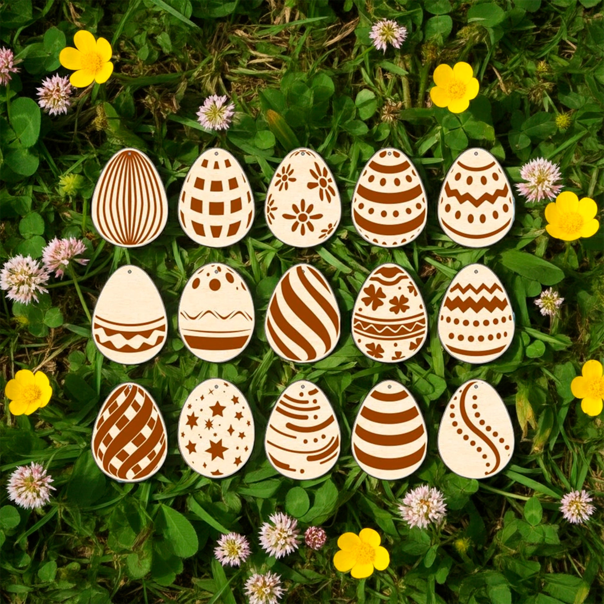 A set of fifteen light brown laser cut wooden plywood Easter egg ornaments, each engraved with a different geometric or floral pattern, lying on a bright green grass background among yellow buttercups and small pink flowers