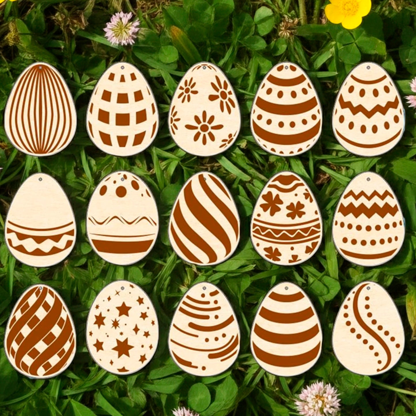 A set of fifteen light brown laser cut wooden plywood Easter egg ornaments, each engraved with a different geometric or floral pattern, lying on a bright green grass background among yellow buttercups and small pink flowers