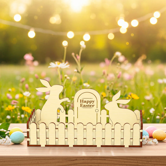 Laser cut  plywood wooden Easter fence decoration featuring two bunny silhouettes and a sign that reads "Happy Easter," styled outdoors on a table with colorful eggs and a wildflower background