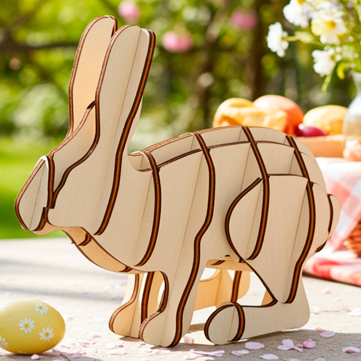 A laser cut wooden plywood Easter bunny decoration, assembled from light wood, stands on a sunlit outdoor table with scattered petals, next to a picnic basket and a bouquet of wildflowers.