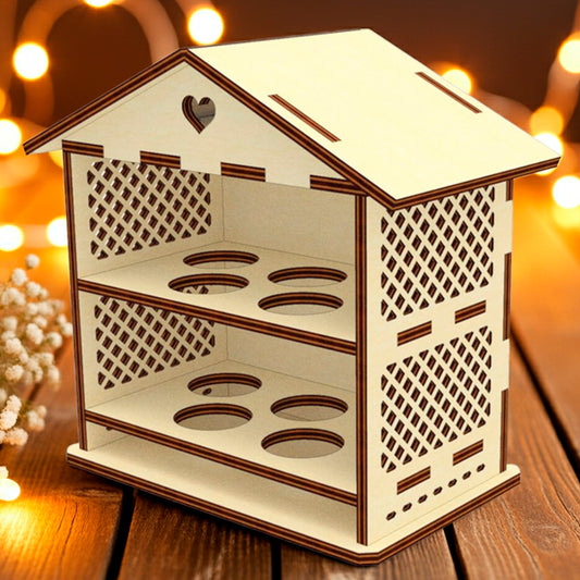 Laser cut plywood wooden house-shaped Easter egg holder with lattice sides and a heart cutout on the roof, featuring two shelves with four round slots each. It rests on a rustic wooden table with baby's breath flowers and warm, blurry fairy lights in the background.