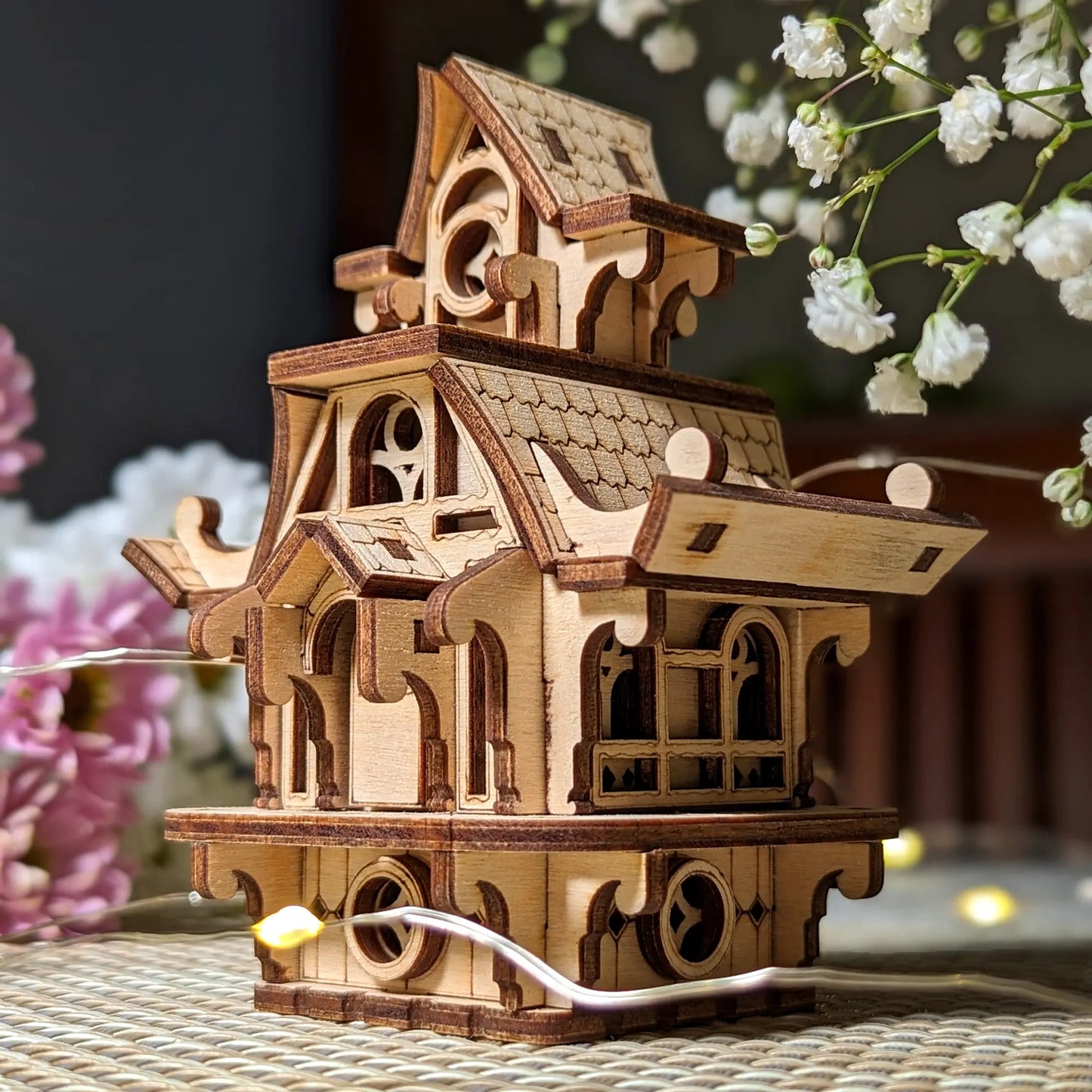 Side angle of a laser cut plywood fairy house. Showcases layered wooden roof structure and ornate window cutouts.