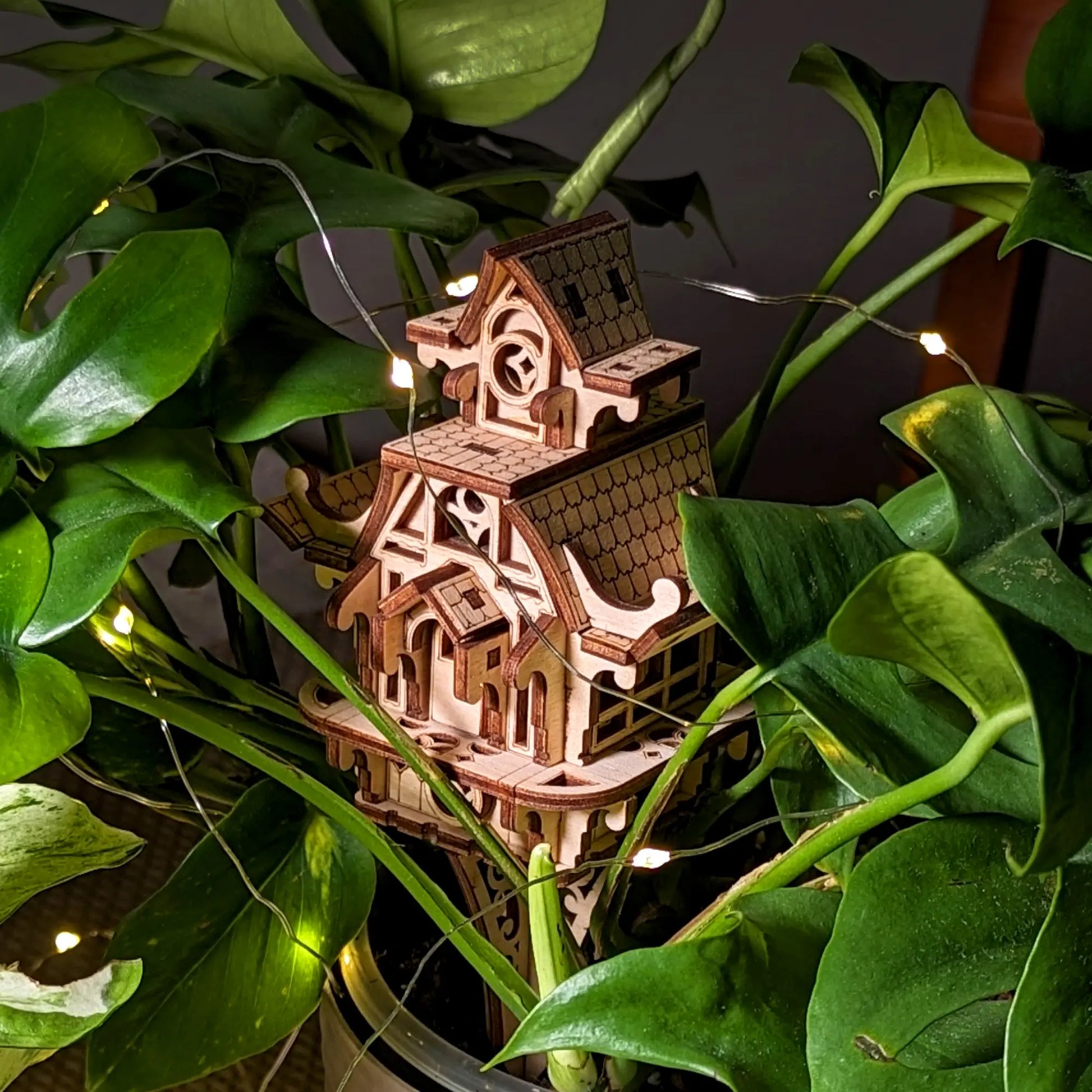 Miniature laser cut plywood fairy house placed inside a lush green plant. Shows scale and detailed wood craftsmanship.