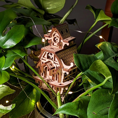 Miniature laser cut plywood fairy house placed inside a lush green plant. Shows scale and detailed wood craftsmanship.