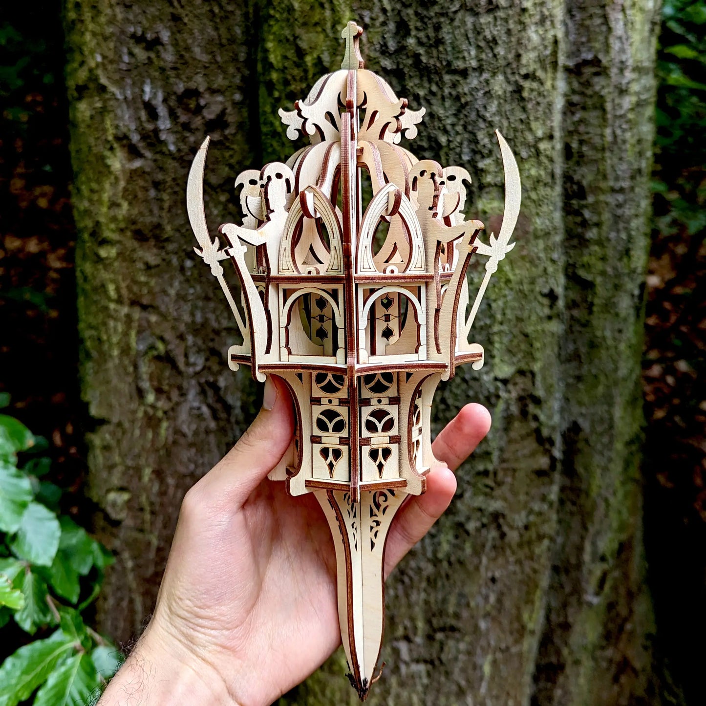A hand holding a miniature fantasy laser cut plywood tower model with curved blades against a blurred forest background.
