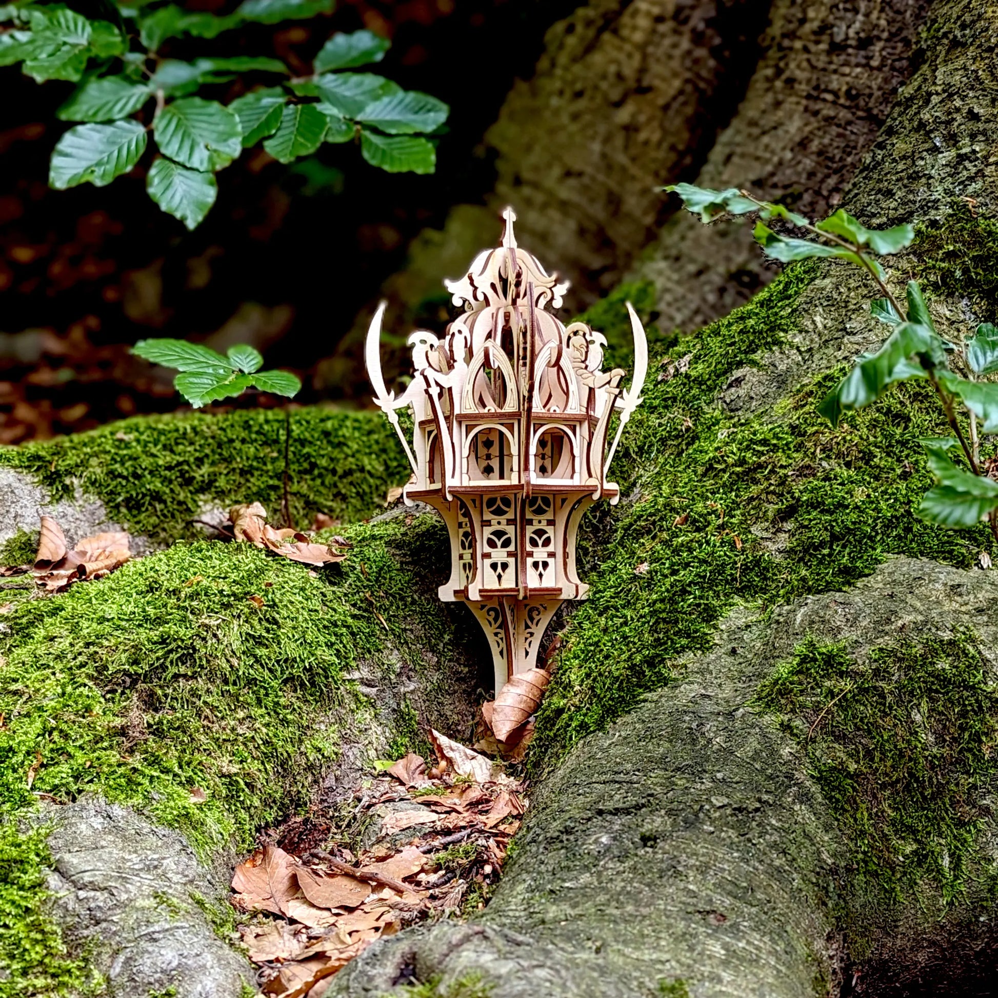 Eye-level view of a decorative laser cut plywood elven shrine miniature with arch windows staged on mossy woodland ground.