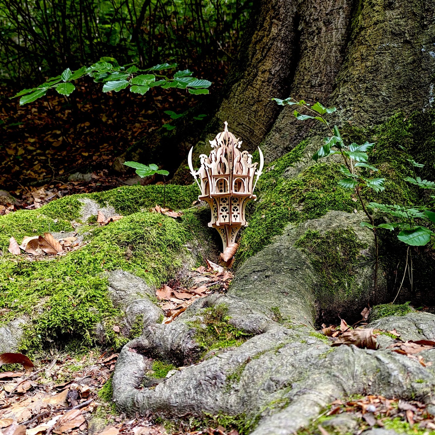 Wide shot of a fantasy laser cut plywood elven tower model placed outdoor among mossy green tree roots in a forest.