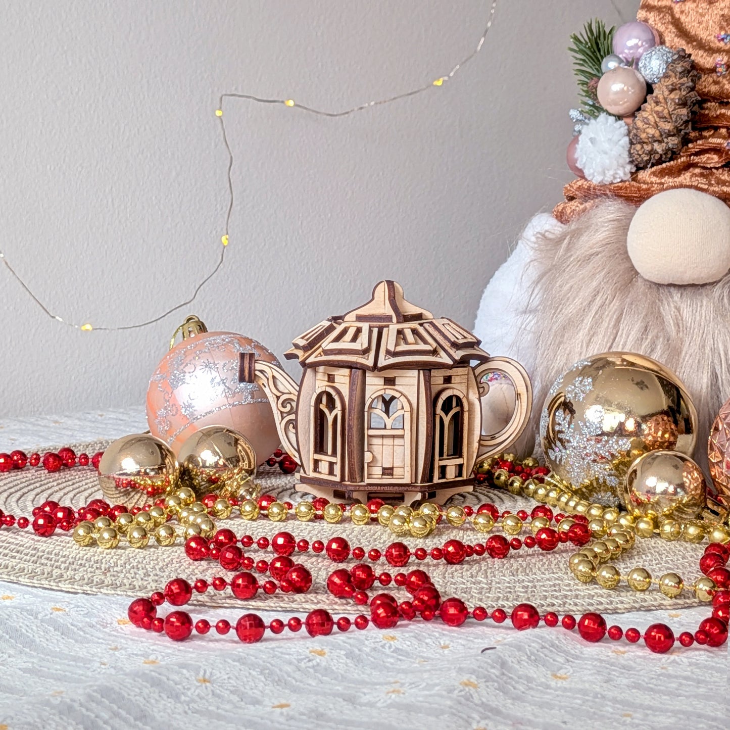 A laser cut wooden teapot fairy house displayed on a festive table with ornaments, red and gold bead garlands, and warm decorative lights
