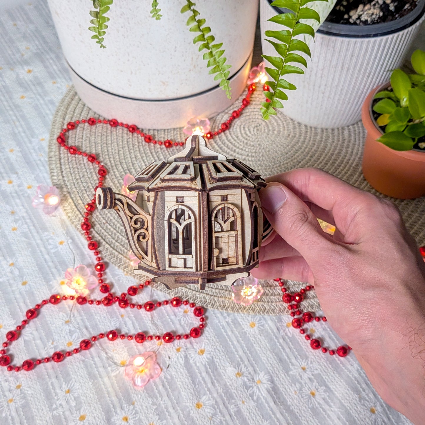 A hand holding the assembled laser cut teapot fairy house near potted plants and festive string lights on a light woven surface