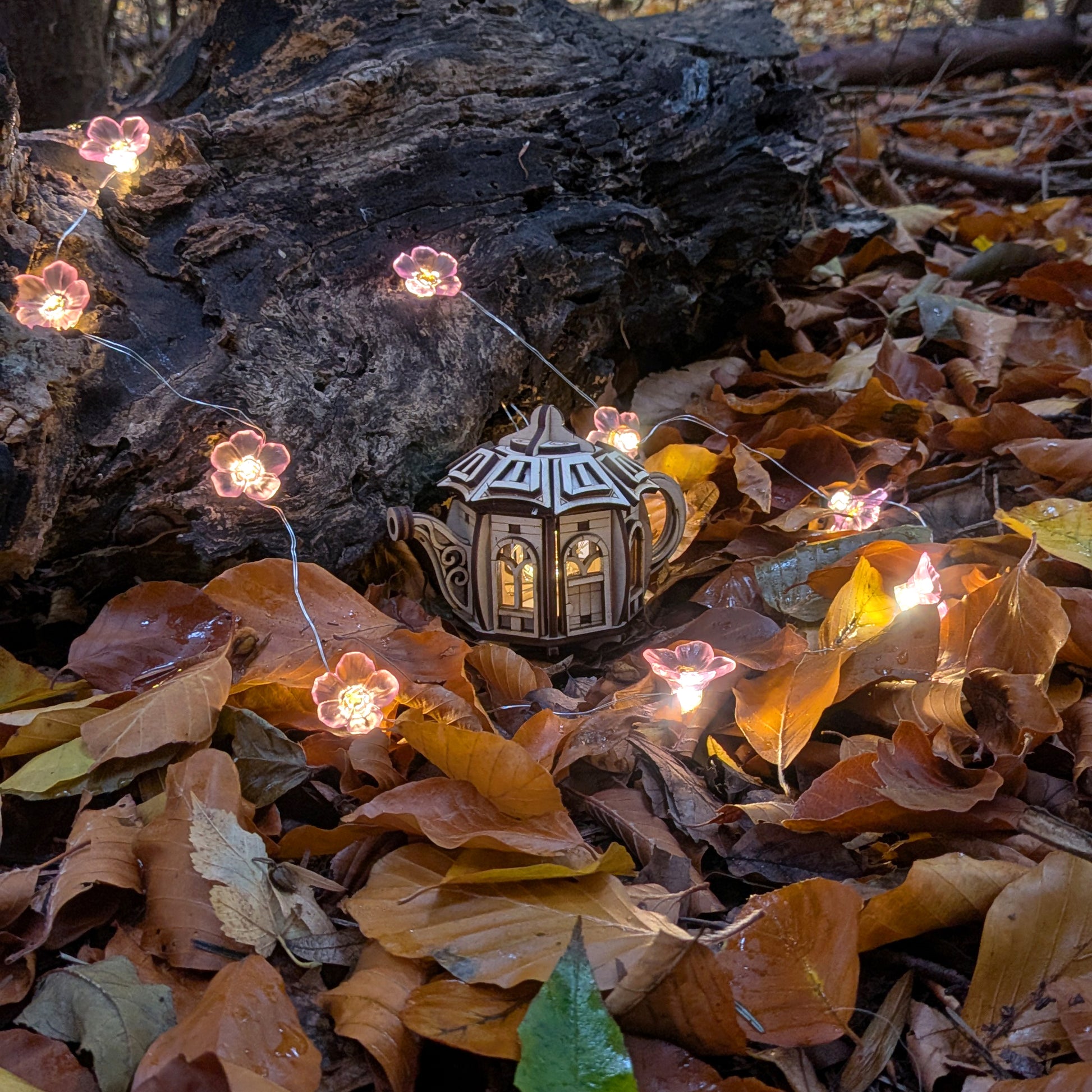 A miniature wooden fairy house shaped like a teapot, made from laser cut plywood, placed among autumn leaves with small glowing flower lights surrounding it.