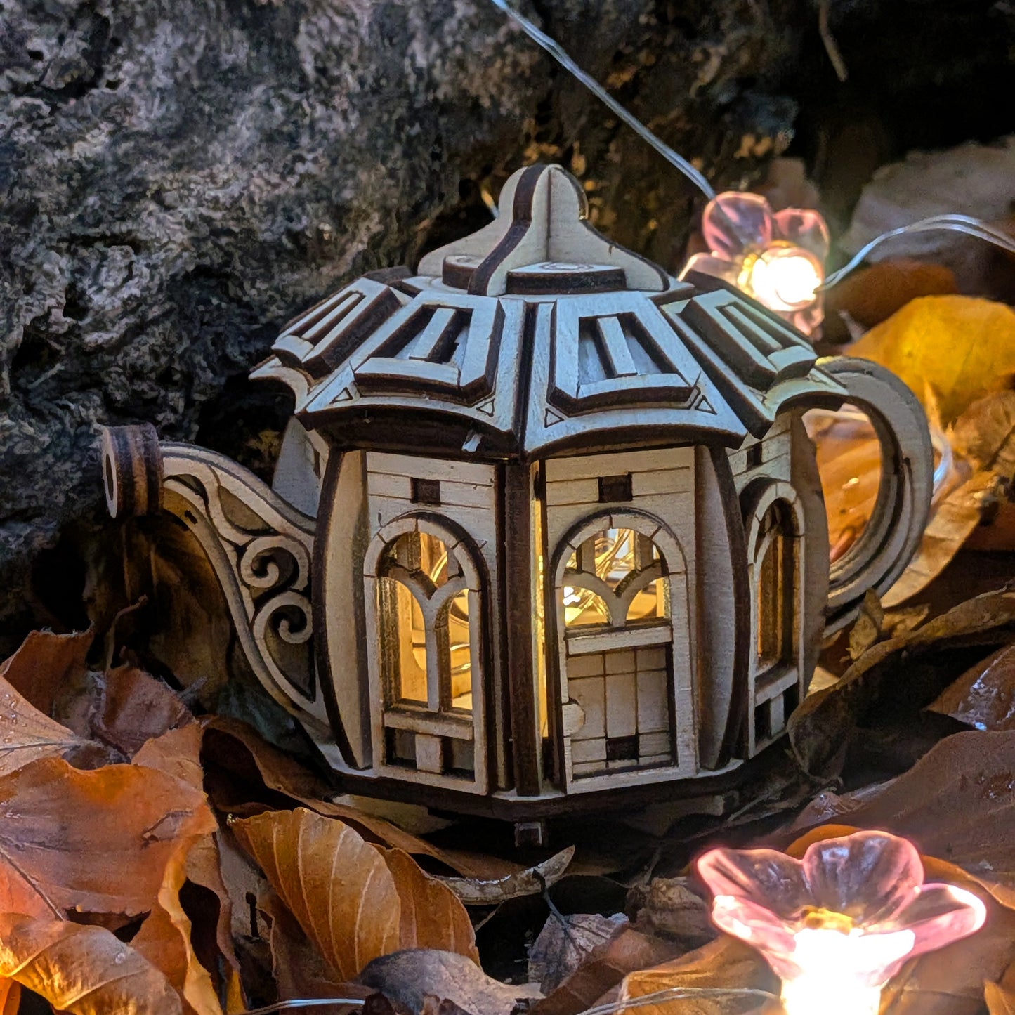 A close-up of the laser cut wooden teapot fairy house showing detailed arched windows and layered plywood structure, illuminated from within