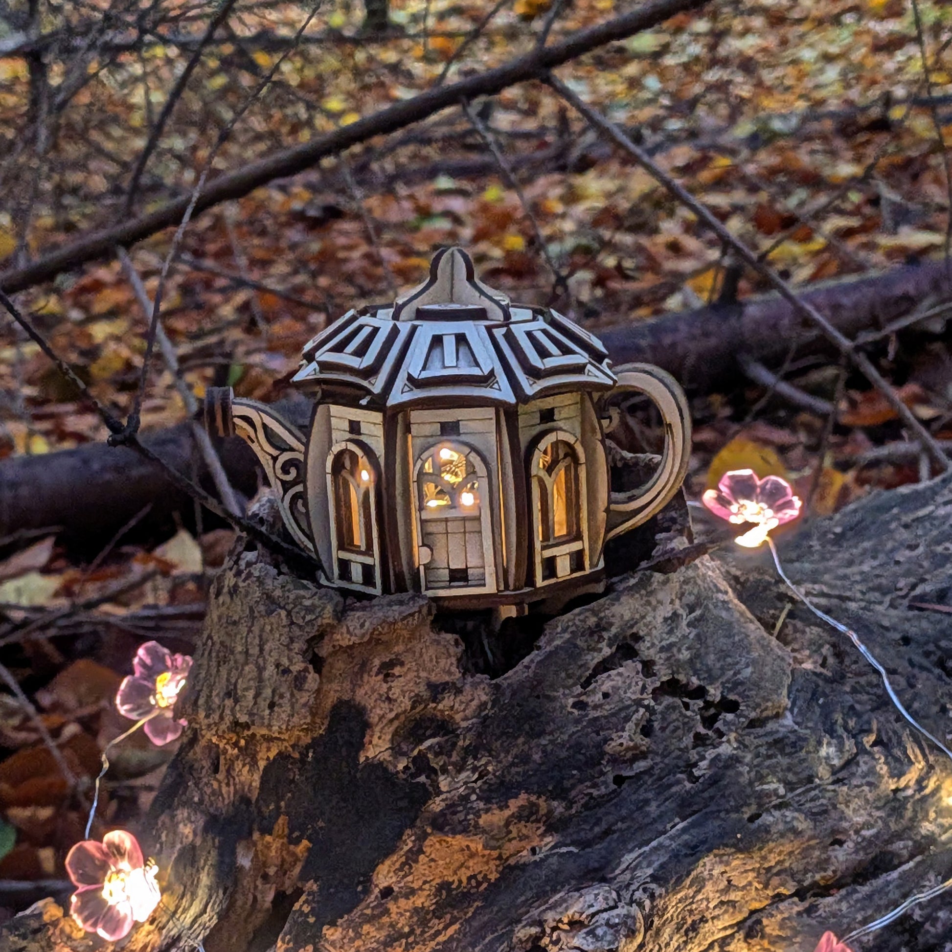 A laser cut plywood teapot-shaped fairy house resting on a tree stump in a forest, softly lit from inside with warm light