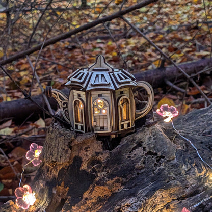 A laser cut plywood teapot-shaped fairy house resting on a tree stump in a forest, softly lit from inside with warm light