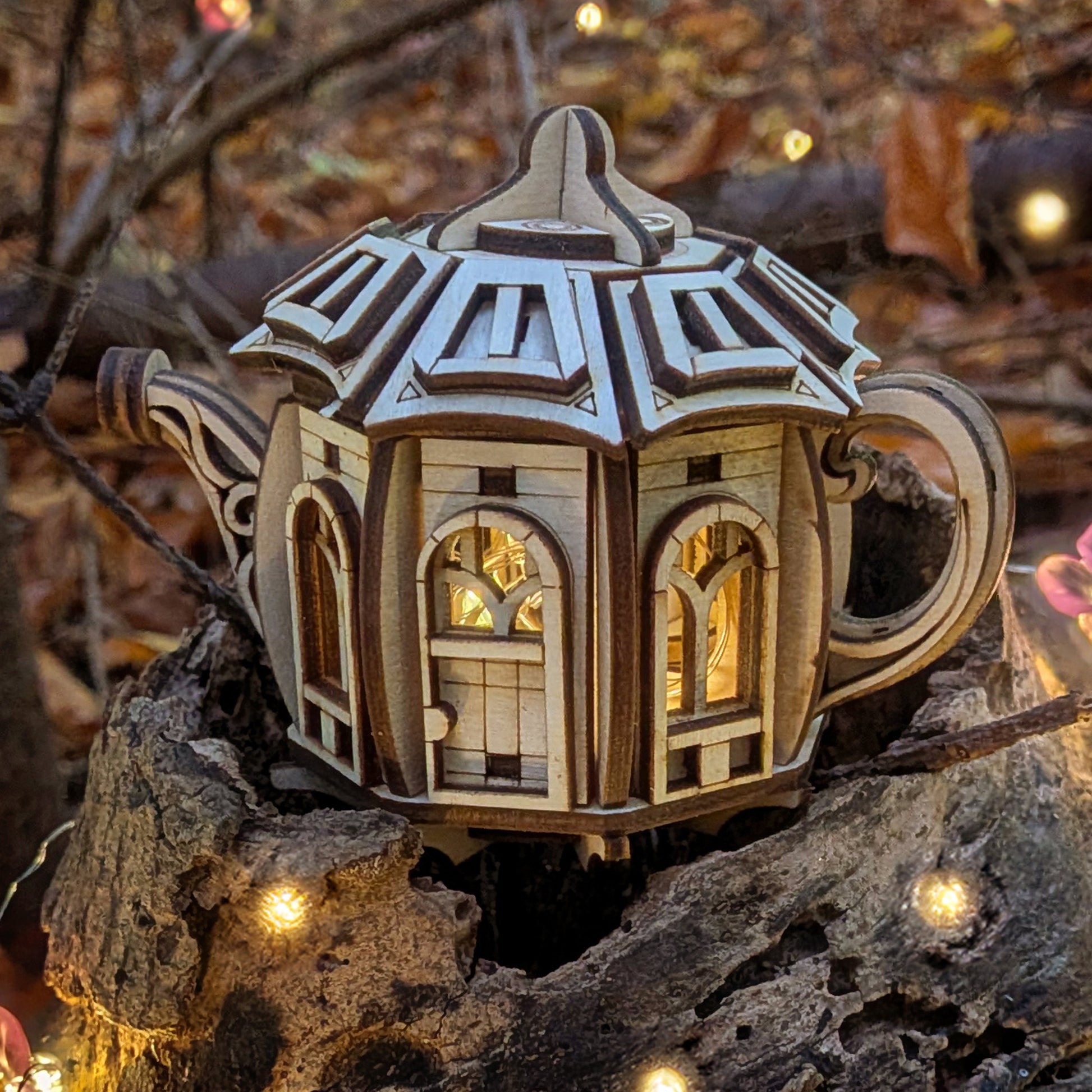 Laser cut wooden fairy house shaped like a kettle, with arched windows glowing from inside, displayed outdoors among tree bark and small decorative lights.