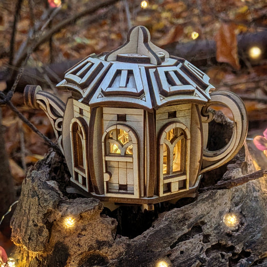 Laser cut wooden fairy house shaped like a kettle, with arched windows glowing from inside, displayed outdoors among tree bark and small decorative lights.