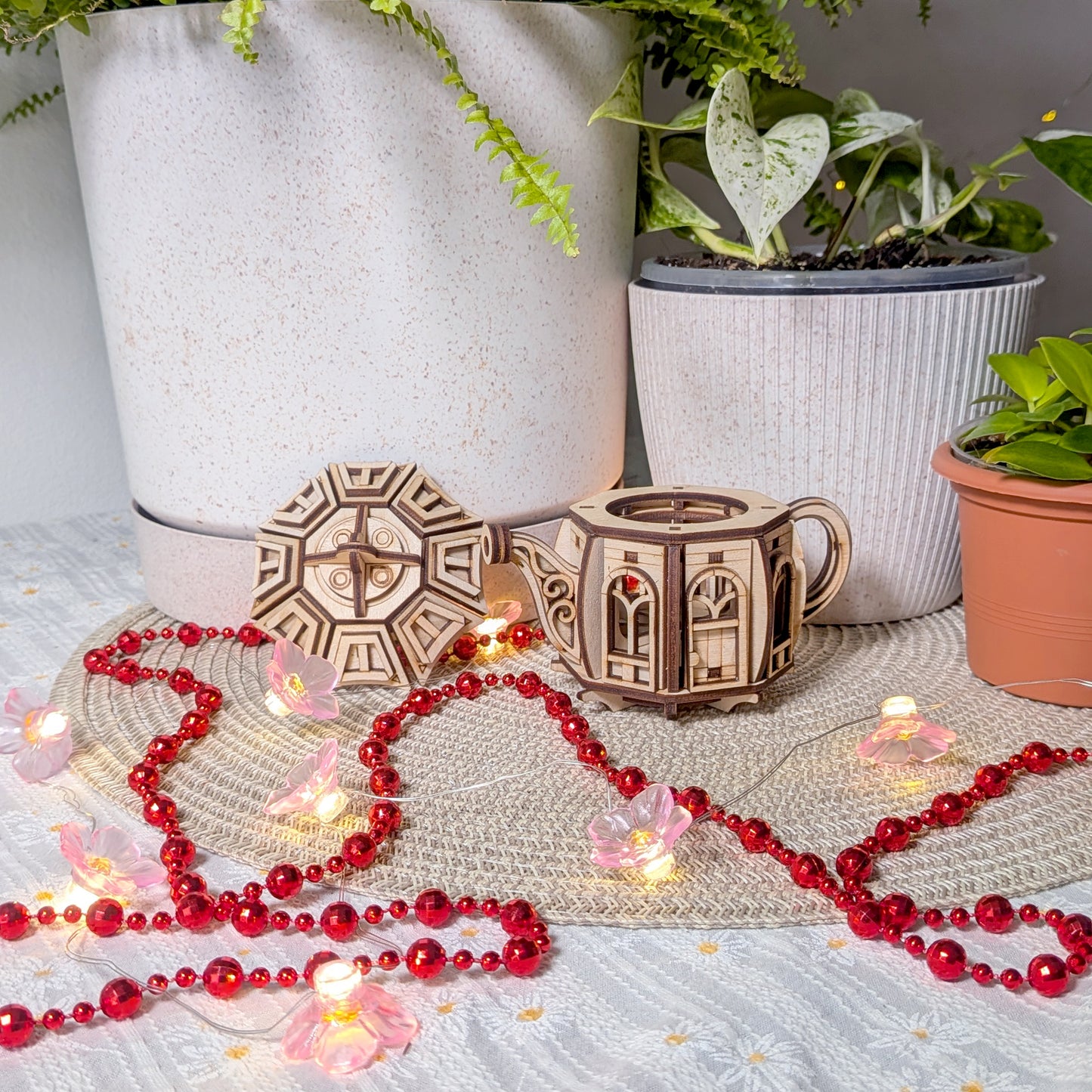 The laser cut teapot fairy house with its removable lid taken off, displayed on a woven mat with potted plants and decorative lights around it.