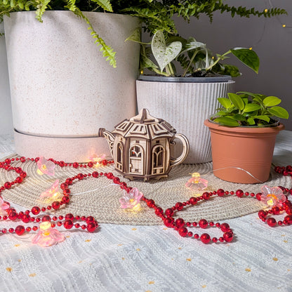 The laser cut wooden teapot fairy house placed on a table among houseplants and pink flower lights, creating a cozy indoor scene