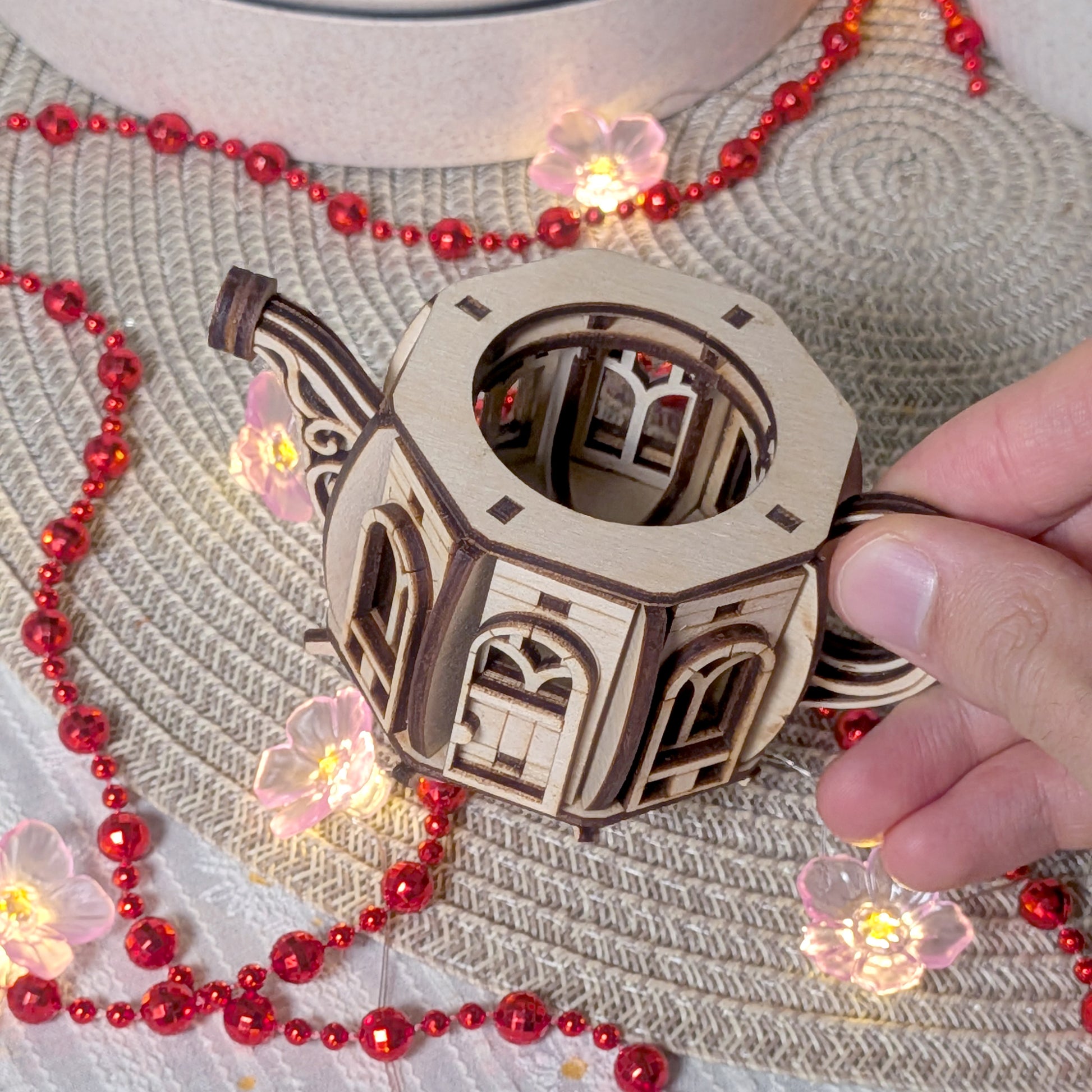 A hand holding the laser cut wooden teapot fairy house body, showing the open top and interior space among decorative lights