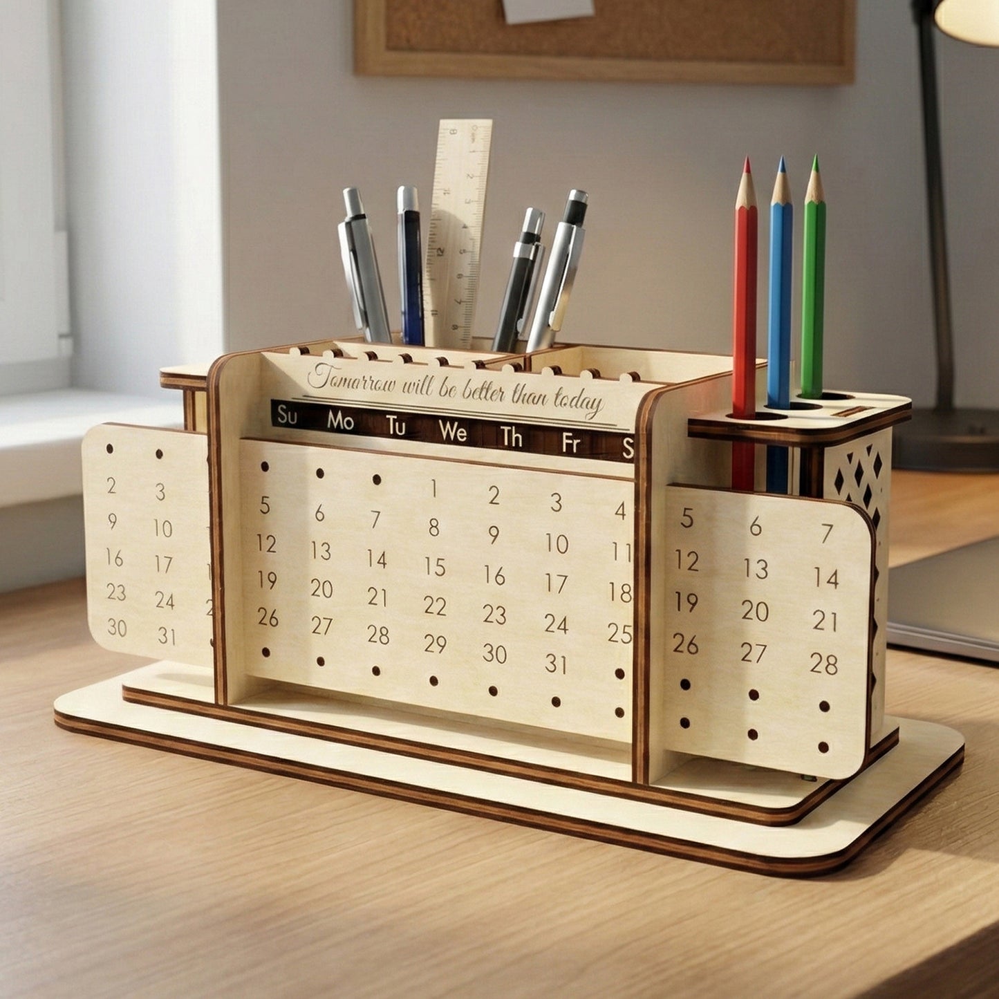 Laser cut plywood desk organizer featuring a perpetual calendar with sliding date panels. The unit includes holders for pens, pencils, and a ruler. It sits on a wooden desk near a laptop and lamp, showing the natural wood grain and dark burnt edges.