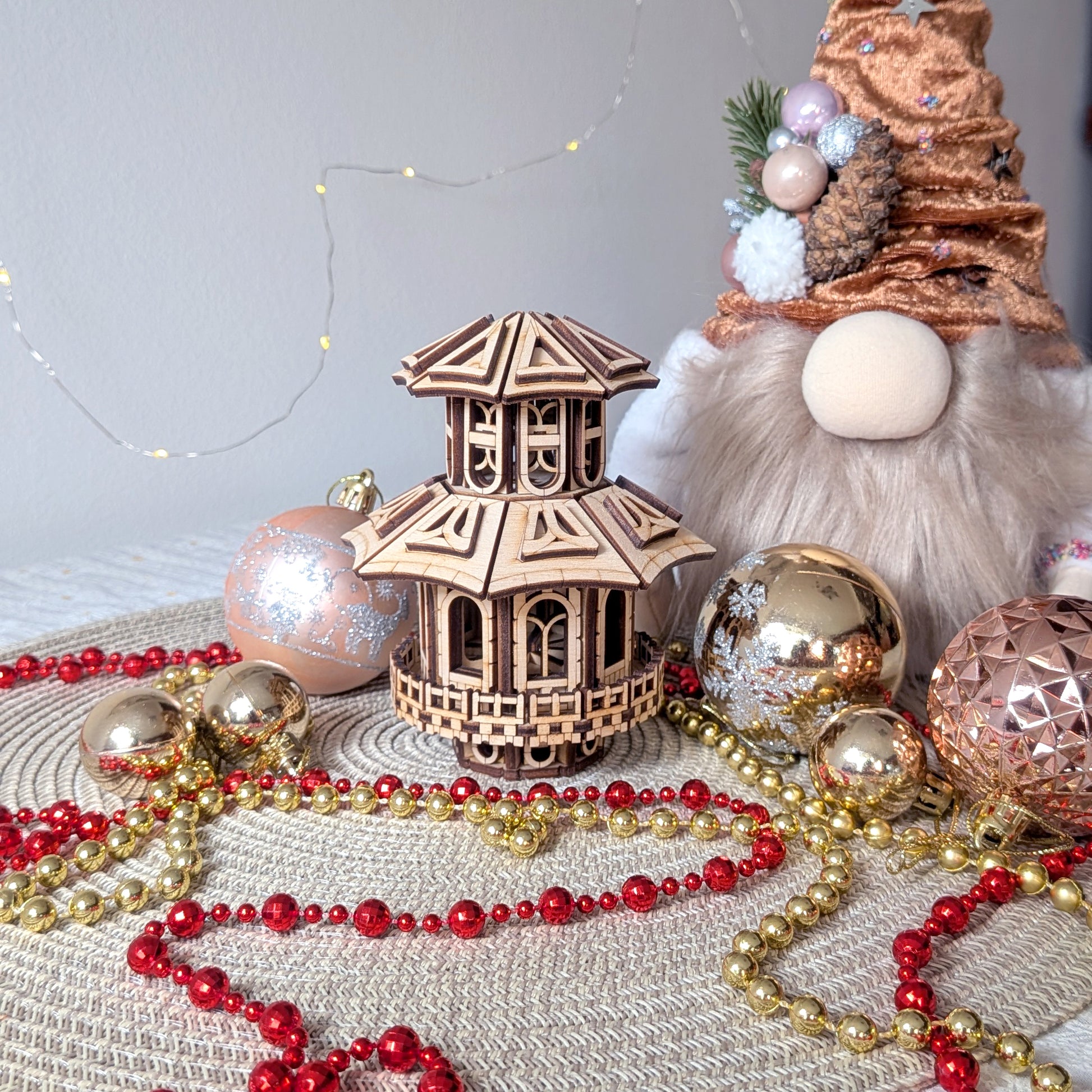 Laser-cut wooden Faelight House surrounded by Christmas decorations, gold and red beads, ornaments, and a holiday gnome in the background