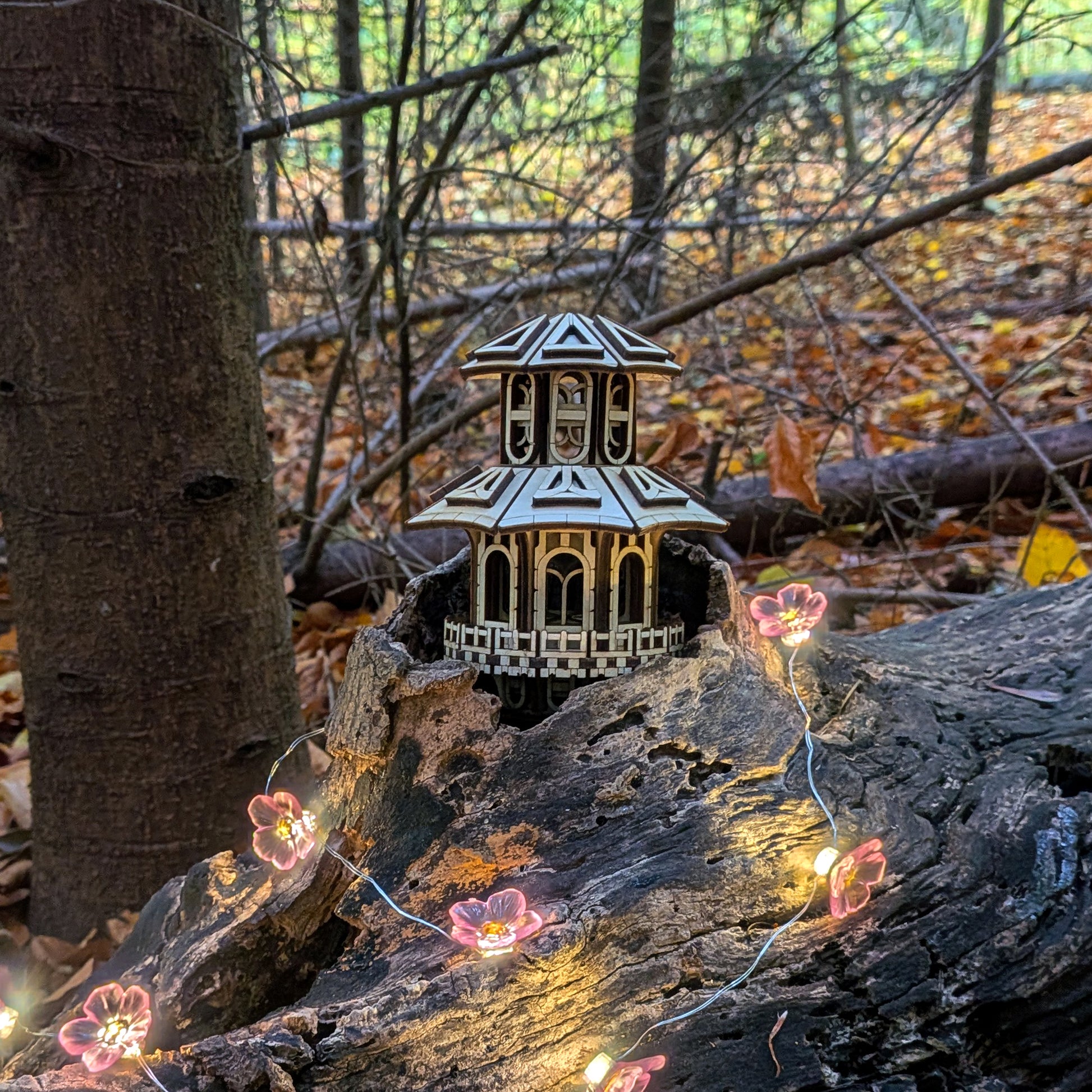 Small laser-cut wooden Faelight House with intricate windows and tiered roof, displayed in the woods with pink flower string lights glowing around it.