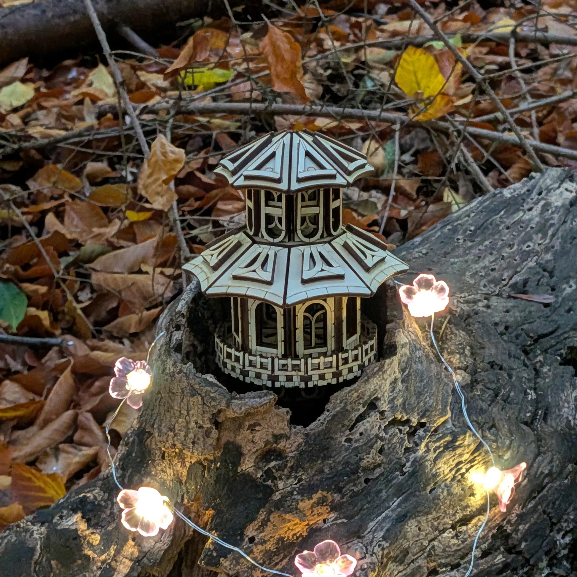 High-angle view of a miniature fantasy fairy house of laser cut plywood nestled inside a mossy tree log with flower lights