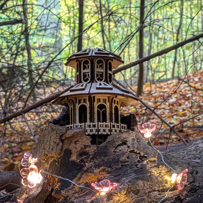 A two-story miniature wooden fairy house with arched windows and layered roof, placed on a tree trunk in a forest surrounded by autumn leaves and warm lights.