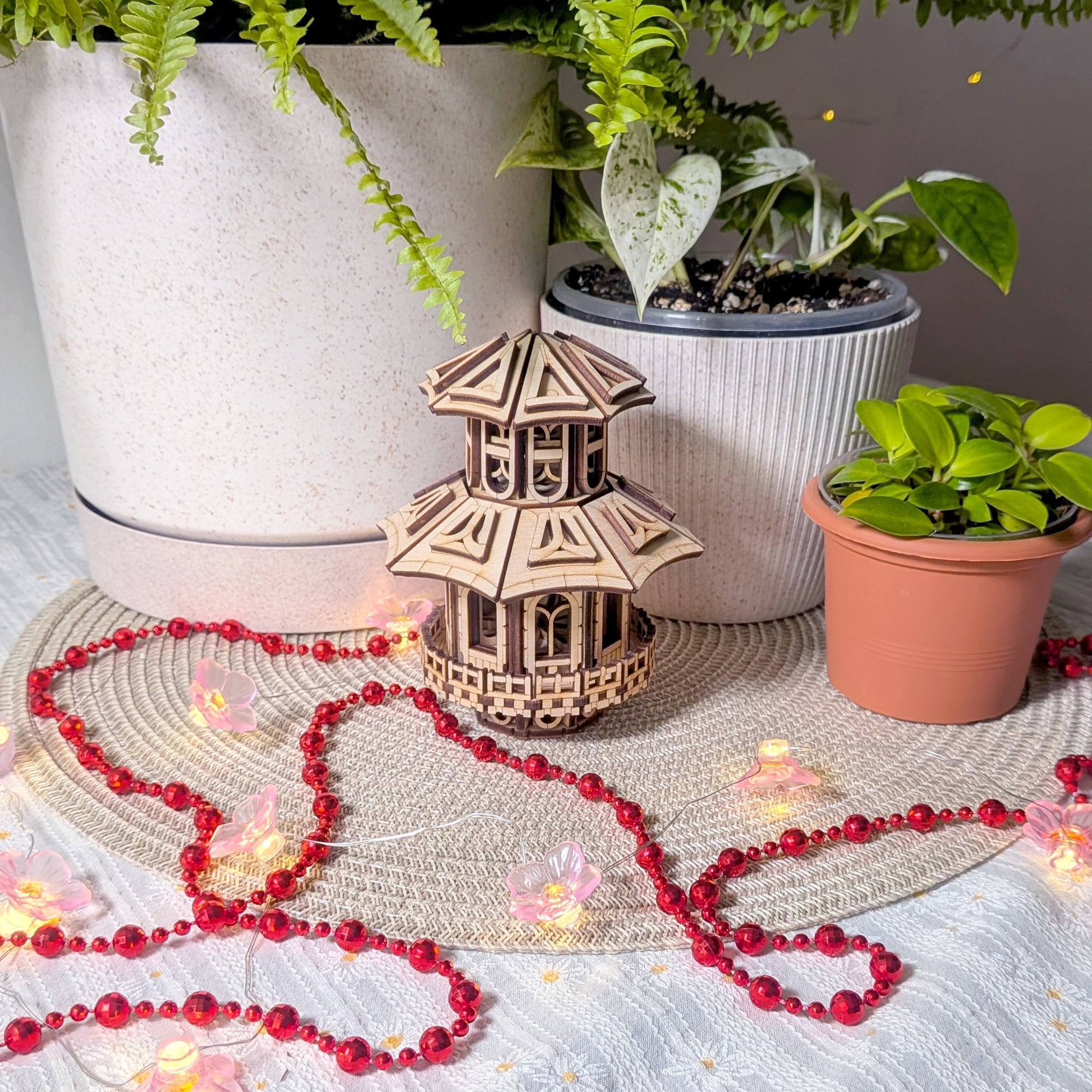 Miniature wooden Faelight House placed beside indoor plants and surrounded by red beads and glowing fairy lights on a woven mat