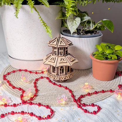 An elegant miniature multi-tier pagoda house model made of laser cut plywood, staged indoors with red beads and house plants