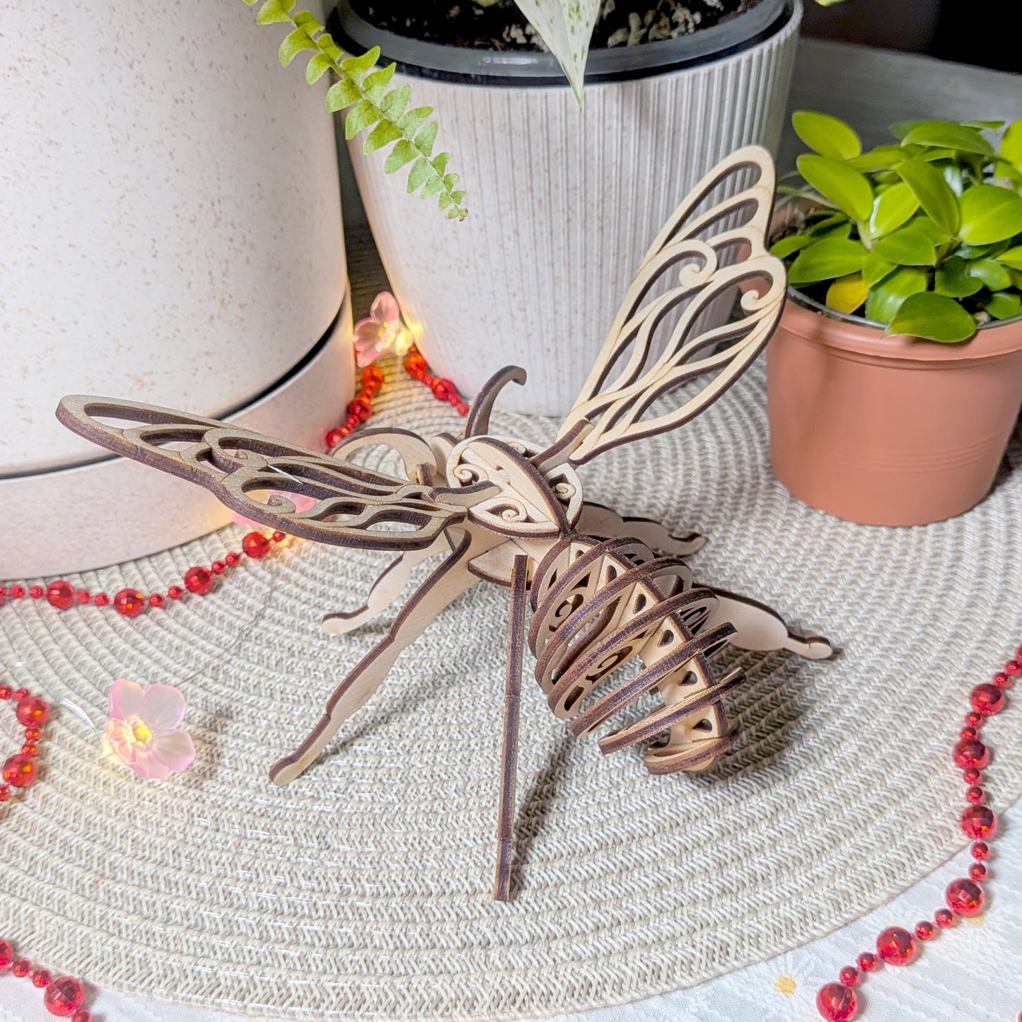 Laser cut fairy bee ornament made from layered plywood, showing detailed body and wings, displayed on a woven mat near potted plants and red lights