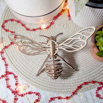Top view of a fairy bee ornament made from laser cut plywood, showing detailed layered wings and body, placed on a woven mat with red beads and plants
