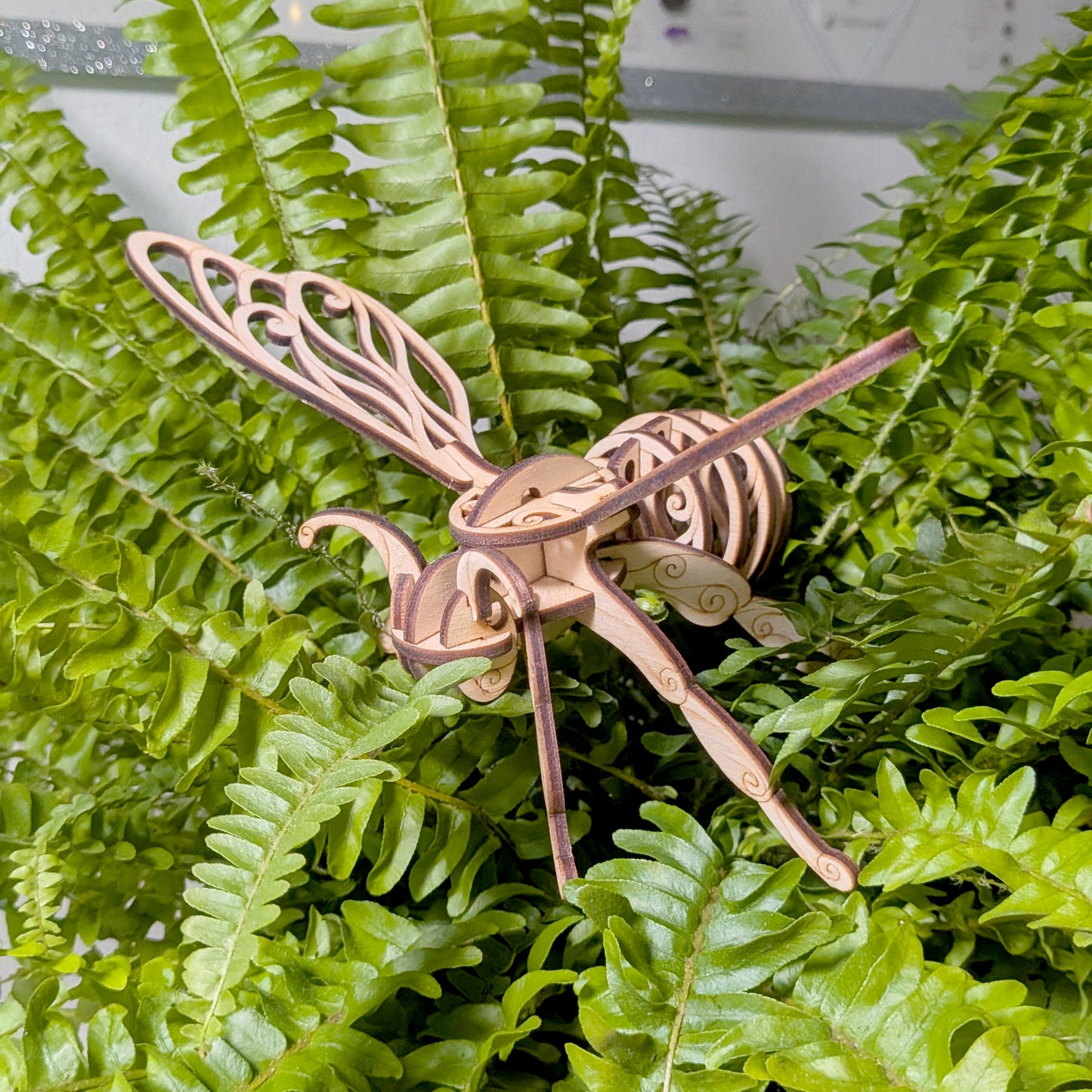 Side view of a laser cut wooden fairy bee ornament displayed on vibrant fern foliage, emphasizing detailed design and natural texture
