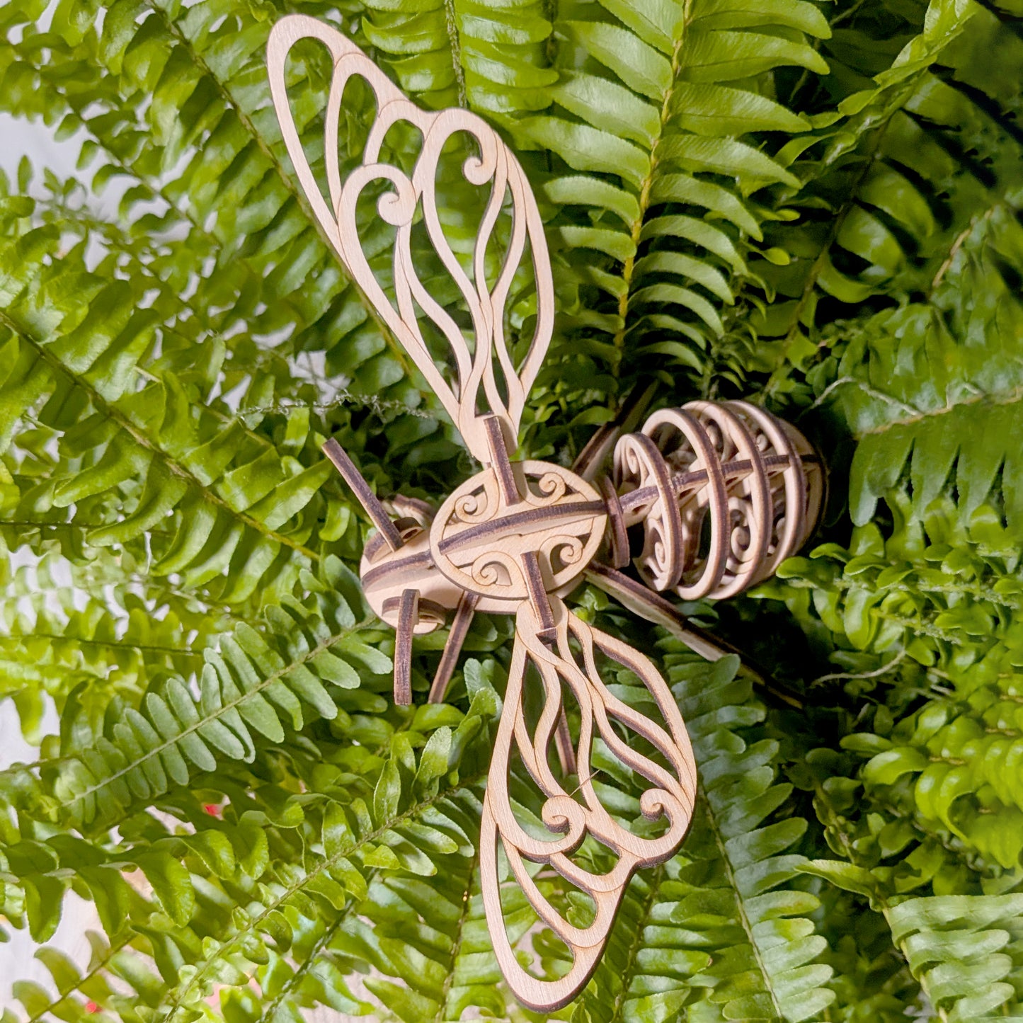 Laser cut fairy bee ornament resting on lush green fern leaves, showing decorative wings and layered wooden construction