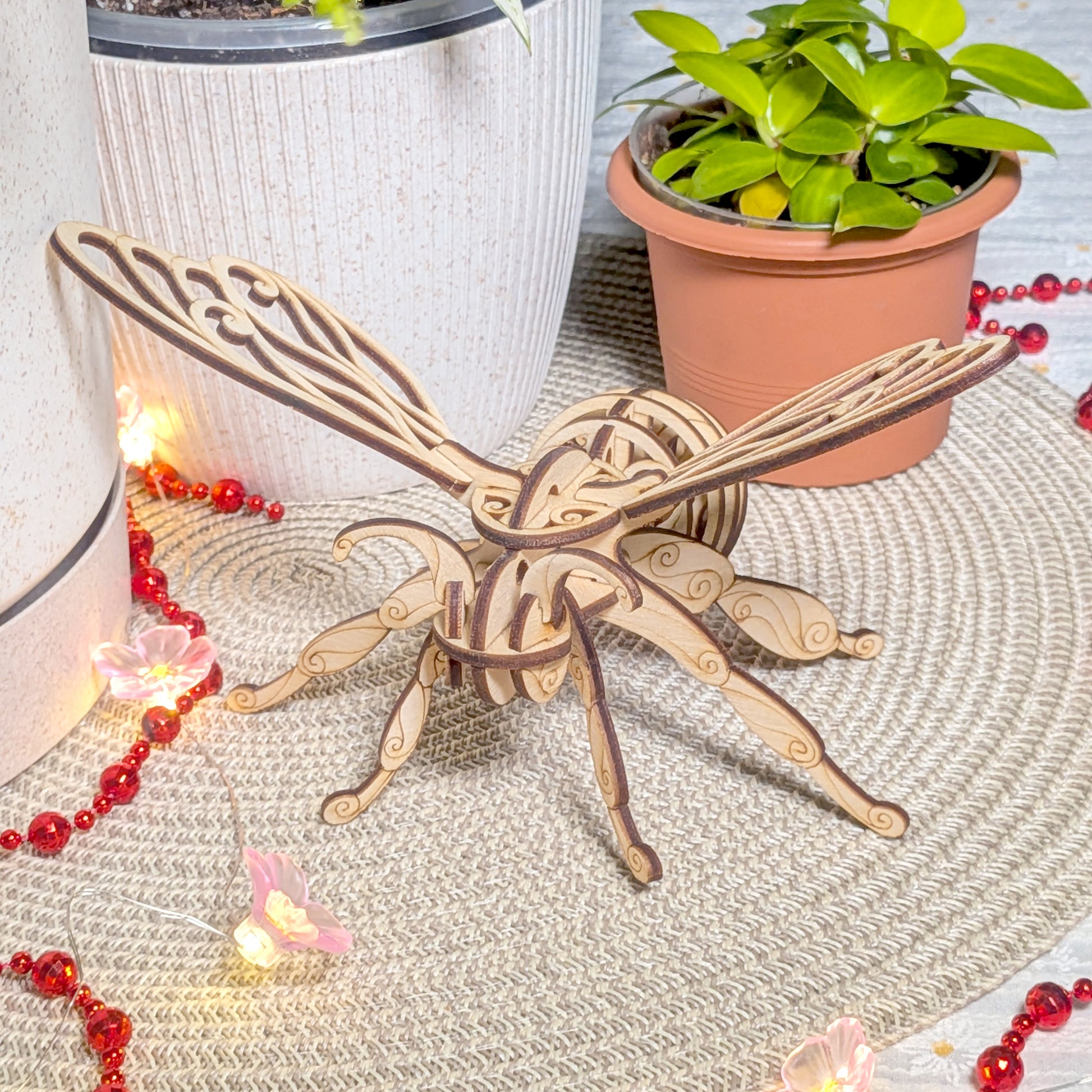 Laser cut wooden fairy bee ornament with decorative wings and curved details, displayed on a round mat surrounded by glowing red beads
